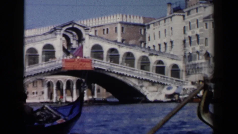 A handheld 8mm view of the Rialto Bridge from a gondola, with blue water, arched stonework, and boat details visible.