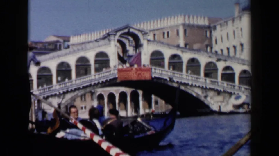 1960 Venice gondola view with the Rialto Bridge, arched bridge shops, canal water, and nearby passengers visible.