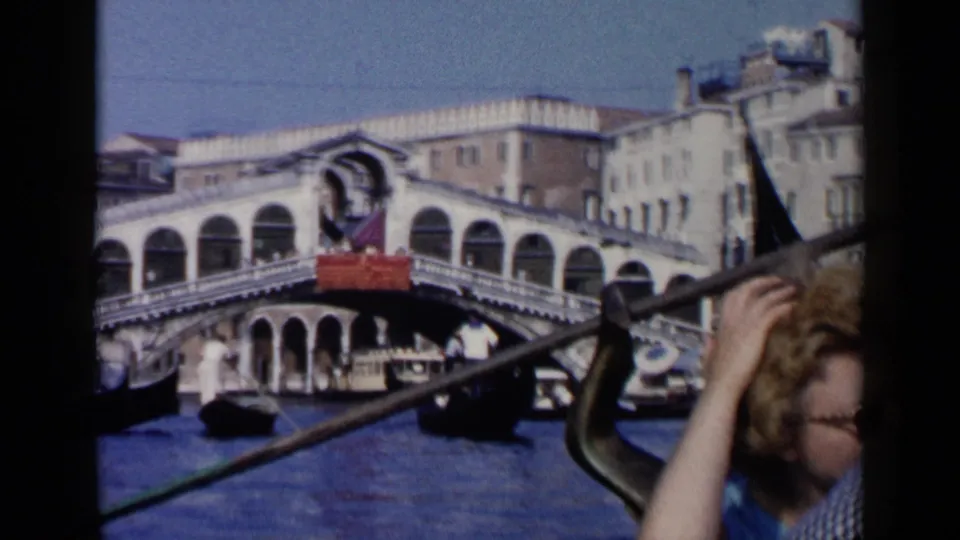 1960 Venice canal scene from inside a gondola, showing the Rialto Bridge, gondolier's oar, and a passenger at the edge of frame.