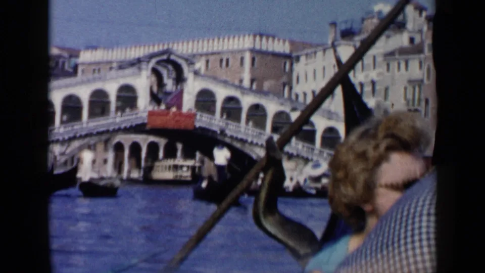 1960 Venice gondola footage with a diagonal oar, a passenger in the foreground, canal water, and the Rialto Bridge in the background.
