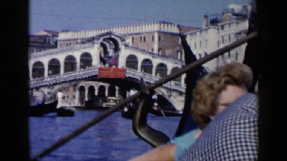 1960 Venice home-movie frame showing a passenger close to camera, a gondola oar, and the Rialto Bridge across the canal.
