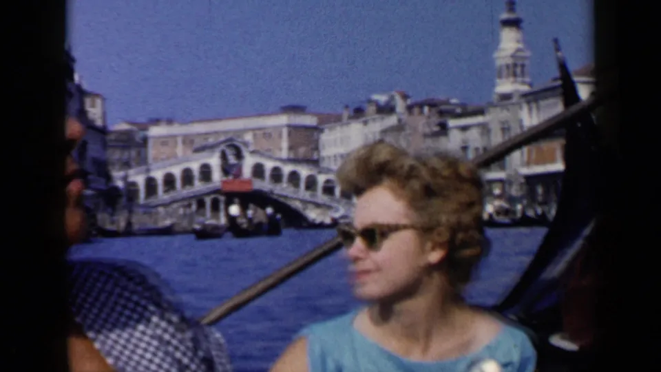 Woman in a blue dress and sunglasses sits in a gondola in Venice, with the Rialto Bridge and canal architecture behind her.