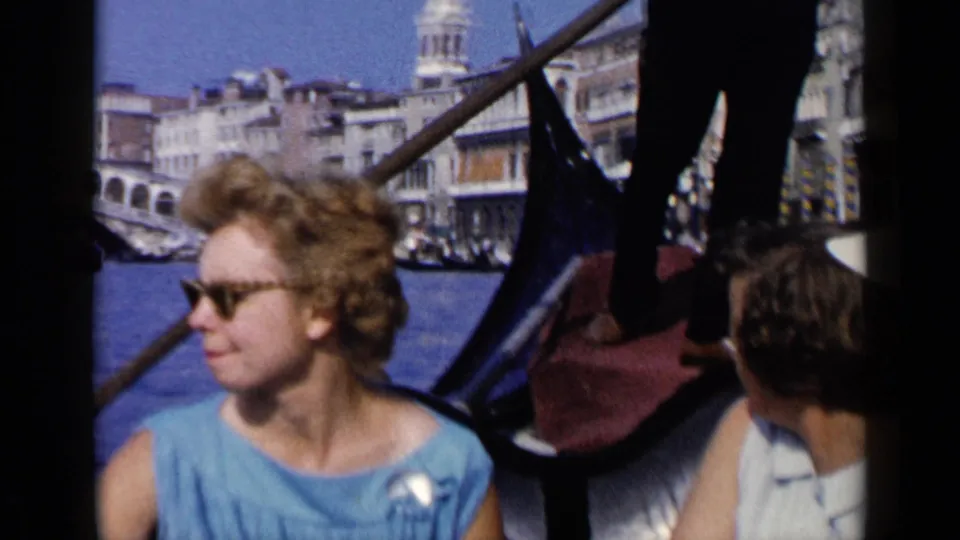 1960 Venice home-movie frame showing a woman in blue seated in a gondola, with passengers, oars, canal water, and city buildings behind her.