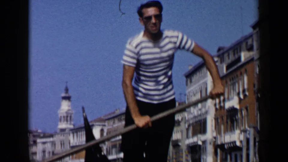 Gondolier in a striped shirt stands with a long oar during a 1960 Venice canal ride, with buildings and blue sky behind him.