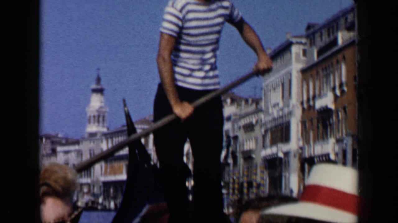 1960 home-movie frame of a gondolier standing with an oar as passengers sit low in a boat on a Venice canal.