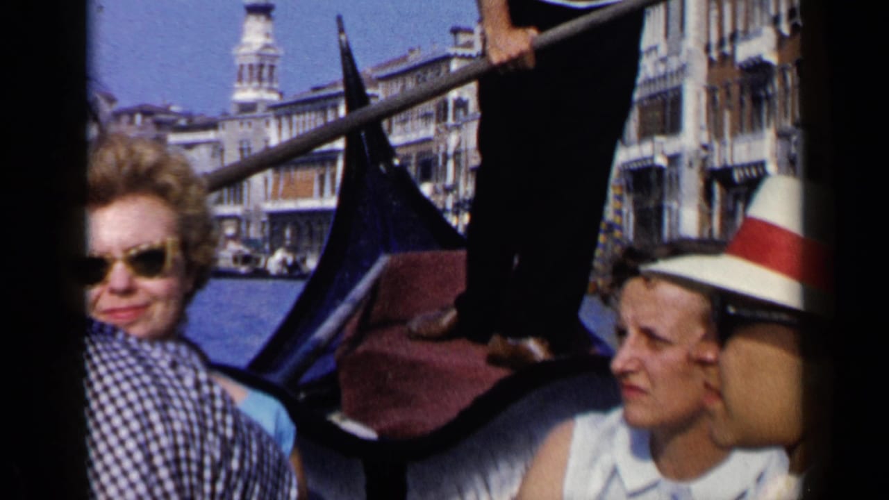 1960 Venice gondola view with two visible passengers, a gondolier's legs and oar, and waterfront buildings behind them.