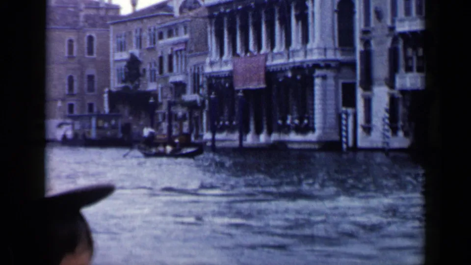 1960 Venice canal view with waterfront architecture, rippling water, and the dark edge of the gondola in the foreground.