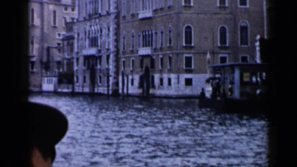 1960 Venice 8mm frame looking across the Grand Canal toward ornate waterfront facades, with a dark frame edge and passenger silhouette in front.