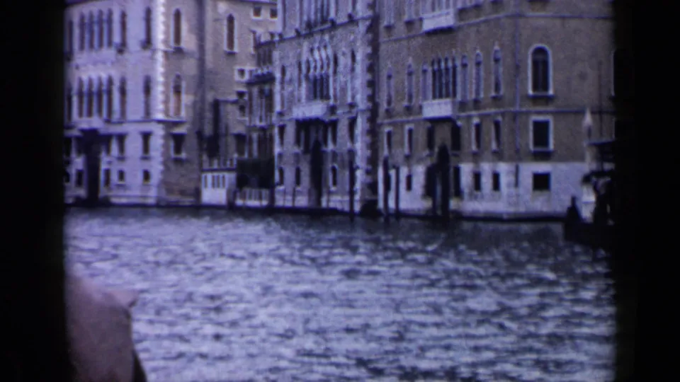 1960 Venice 8mm frame showing canal water, ornate facades, arched windows, and a cropped passenger hat near the lower-left corner.