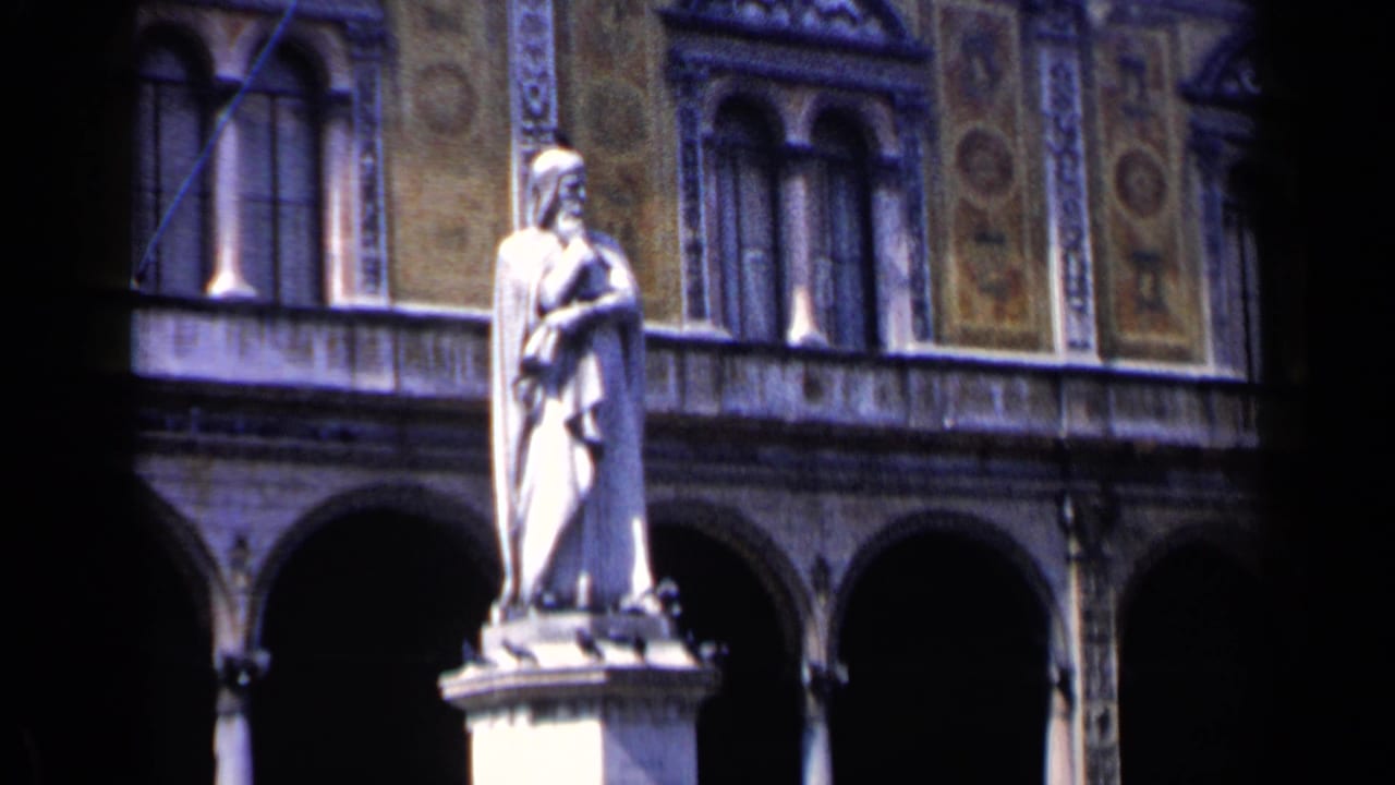 1960 Venice travel footage frame with a robed statue in bright light before dark arches, columns and decorated exterior walls.