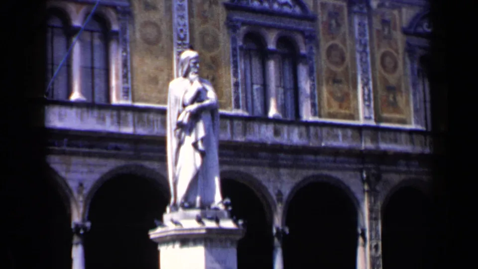 1960 Venice 8mm frame showing a closer view of a pale robed statue on a pedestal before arched courtyard architecture.