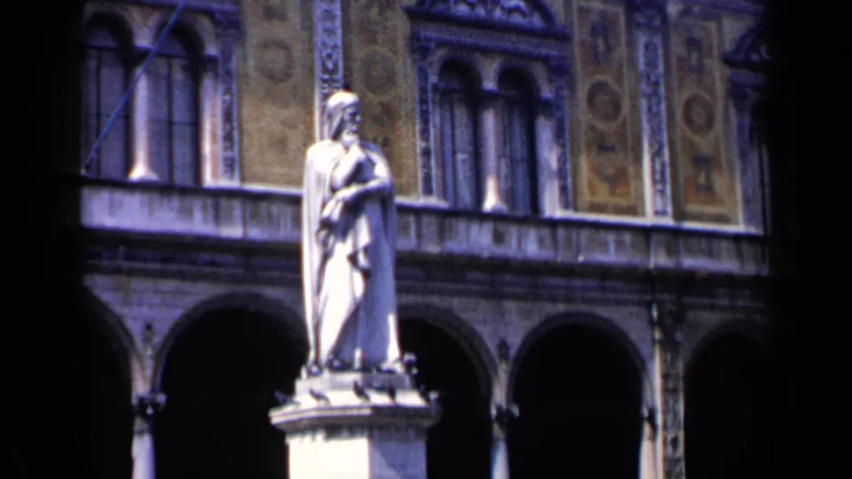 1960 Venice home-movie frame of a robed statue against a historic courtyard facade with arches, columns and patterned panels.
