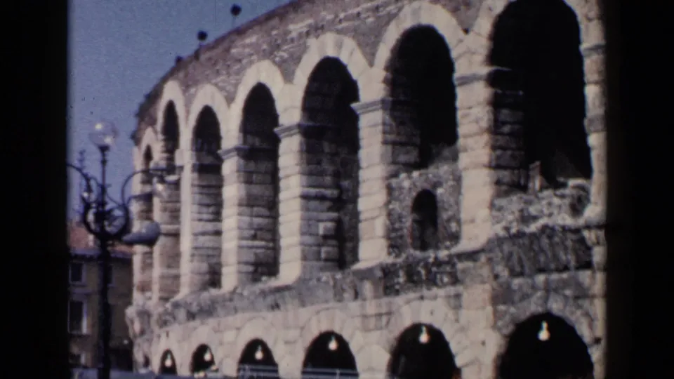 1960 8mm frame showing the arched stone facade of an Italian amphitheater with a streetlamp nearby.