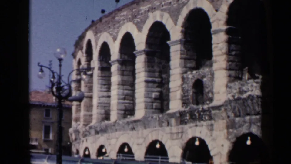 Visitor-shot 1960 8mm view of the amphitheater arches, with nearby buildings and street fixtures at the edge.