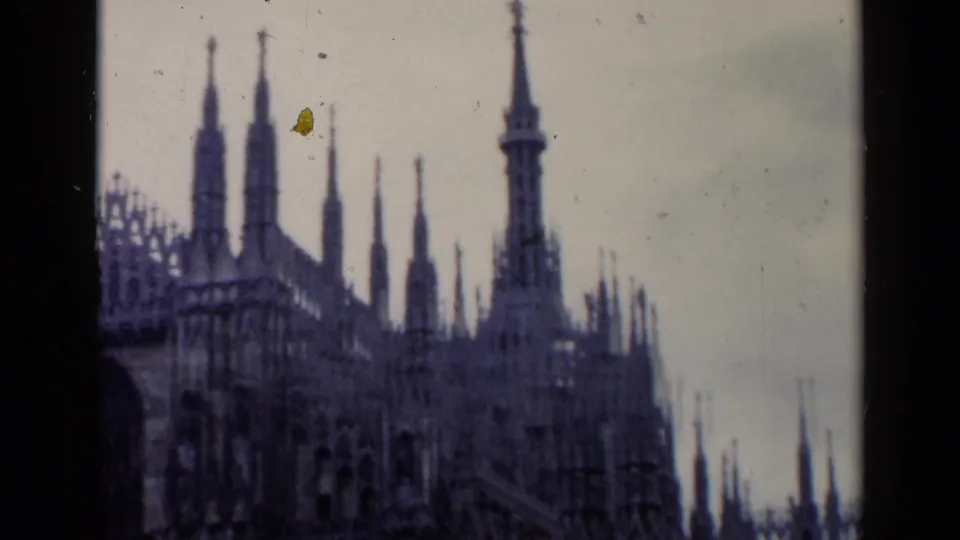 Handheld 1960 travel-film view of an ornate Italian church roofline and spires, framed with grain, dust, and soft focus.