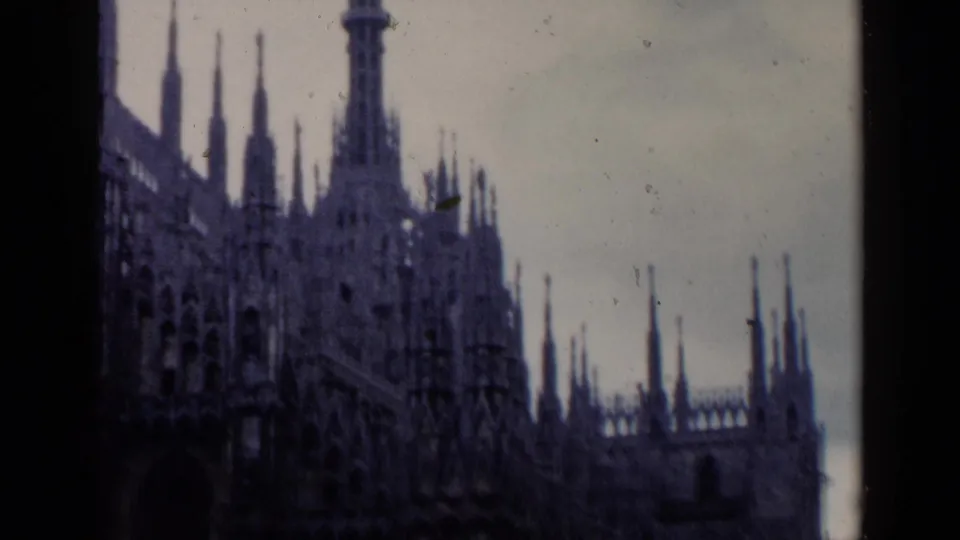 1960 8mm frame of an Italian Gothic church exterior, showing spires, facade ornament, dark frame edge, and visible film grain.
