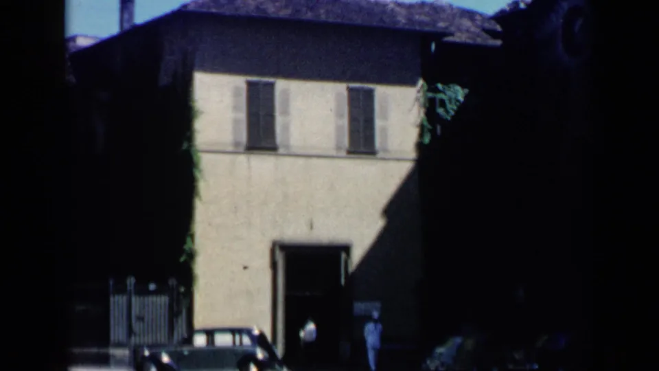 Venice, Italy 1960 frame showing a sunlit two-story facade with shutters, a dark entrance, distant pedestrians, and a small parked car.