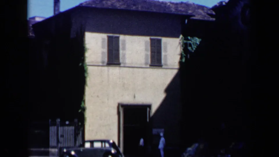 1960 Venice home-movie frame of a pale shuttered building with a gated side area, small car, and distant figures near the doorway.