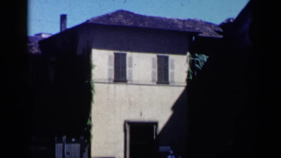 Venice, Italy 1960 frame showing a shuttered building facade with a dark ground-floor entrance, gate, parked car, and strong sunlight.