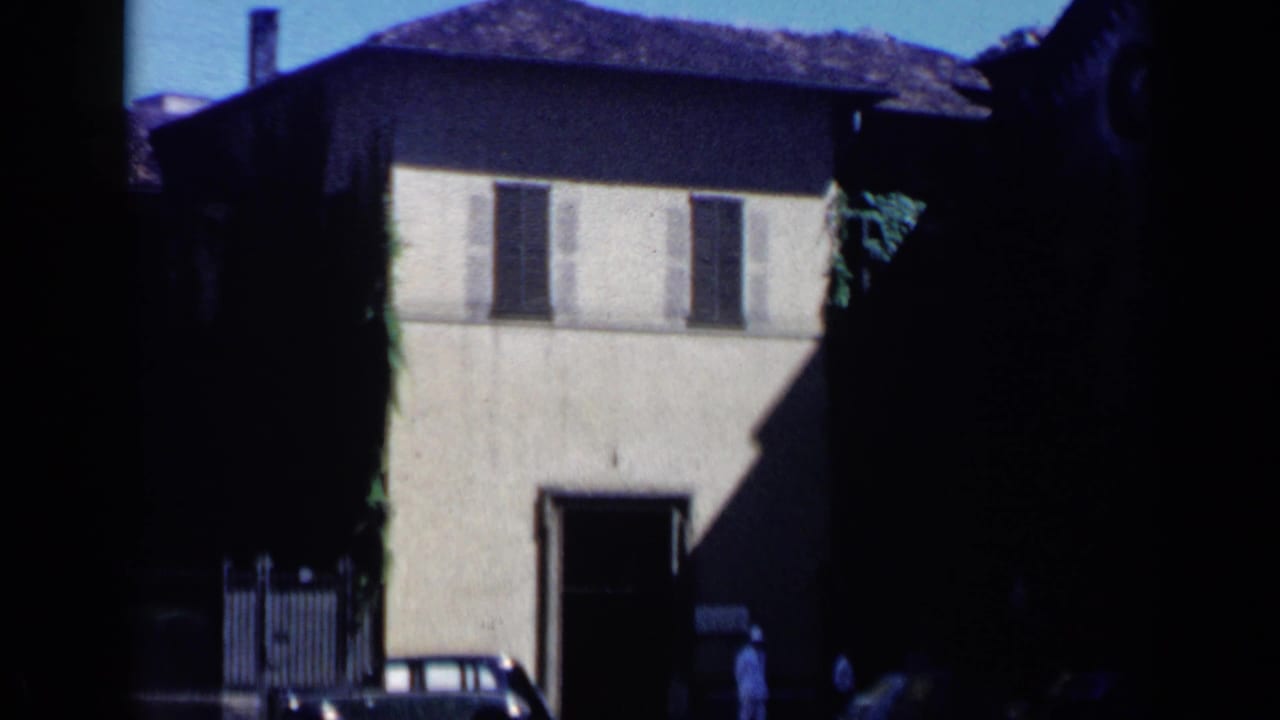 Stucco building with shutters, tiled roof, parked car, and a distant figure near the entrance.
