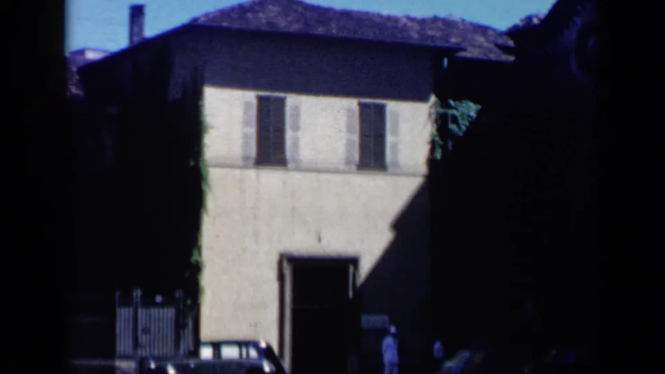 1960 home-movie frame in Italy showing a shaded stucco building, dark shutters, a parked car, and a small figure near an open doorway.