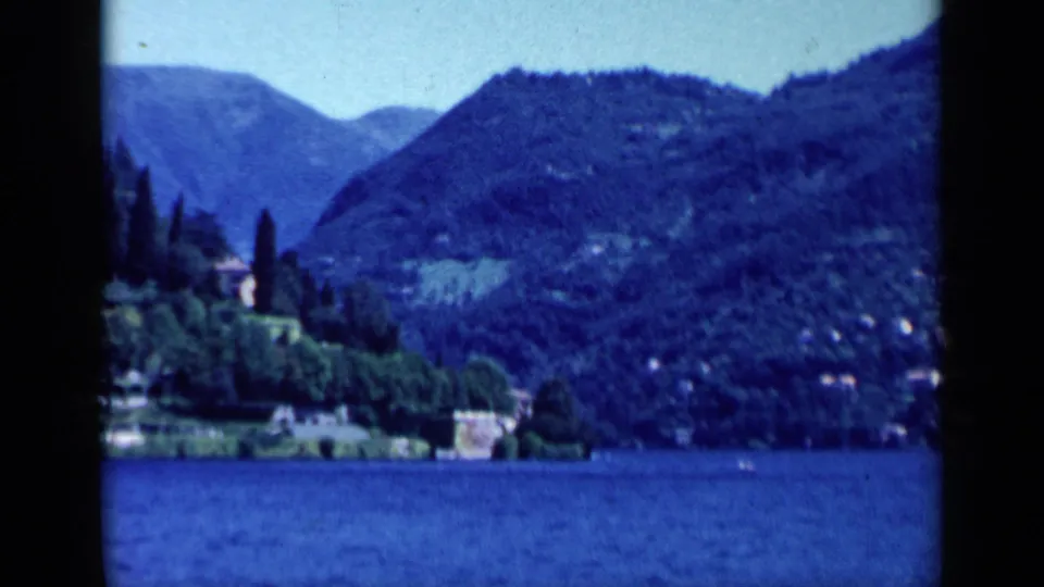 1960 Italy travel footage view of a blue lake, forested hills, shoreline buildings, and tall narrow trees across the water.