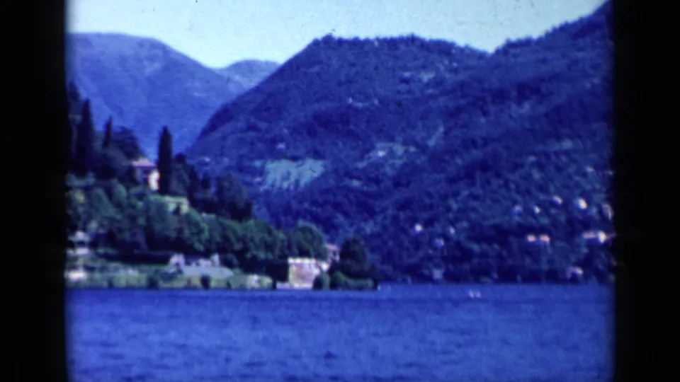Handheld 8mm frame from Italy showing lake water, a tree-covered hillside, shoreline structures, and distant mountain ridges.