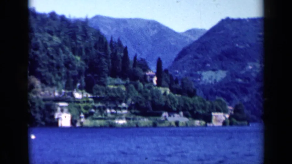1960 Italy lakeside view with shoreline buildings, cypress-like trees, blue water, wooded hills, and mountain ridges in soft 8mm focus.