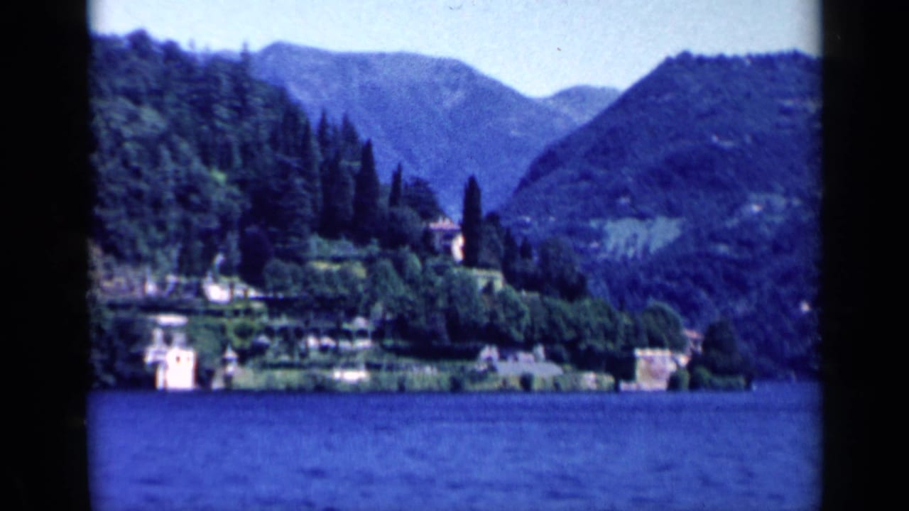 1960 8mm travel view of an Italian lake, wooded mountains, and a distant estate along the shoreline.