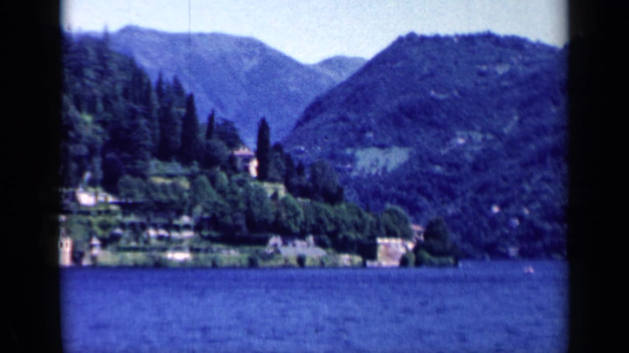 Distant shoreline estate with trees below blue mountain slopes.