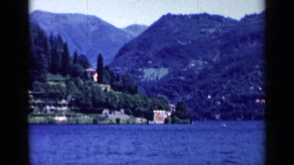 Vintage 8mm lake view in Italy with wooded slopes, shoreline buildings, and soft color shift from the film scan.