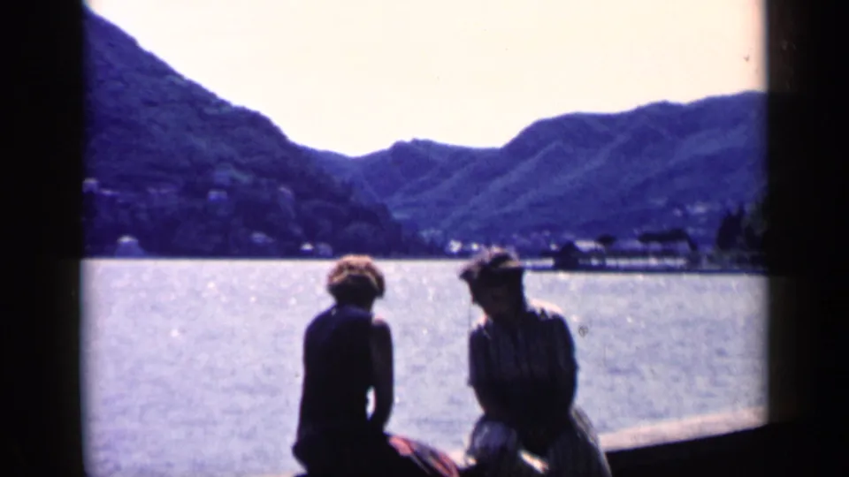 Two seated figures on a lakeside dock look across the water toward mountains in 1960 Italian home-movie footage.