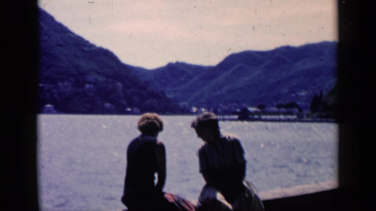 Two adults sit on a ledge beside bright water with hills across the lake.