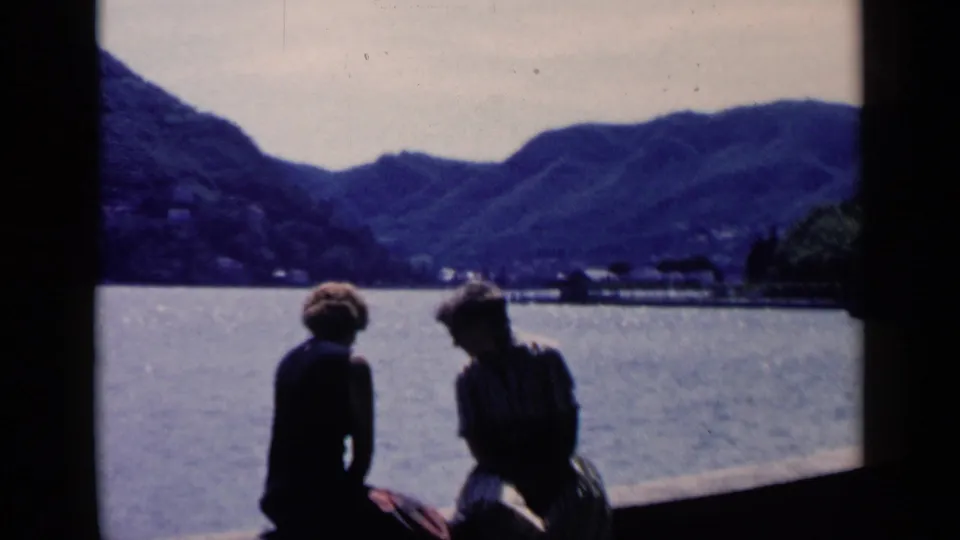 A silhouetted couple sits by a lake in the 1960 Italy footage, framed against bright water and a hilly shoreline.