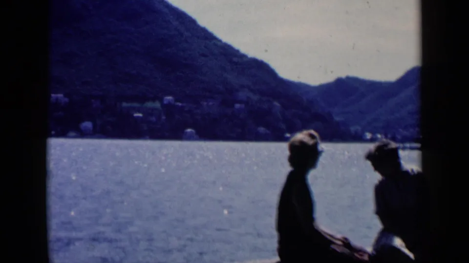 The 8mm frame shows the couple seated by the water, with the woman in a dark top and the man near the right edge.