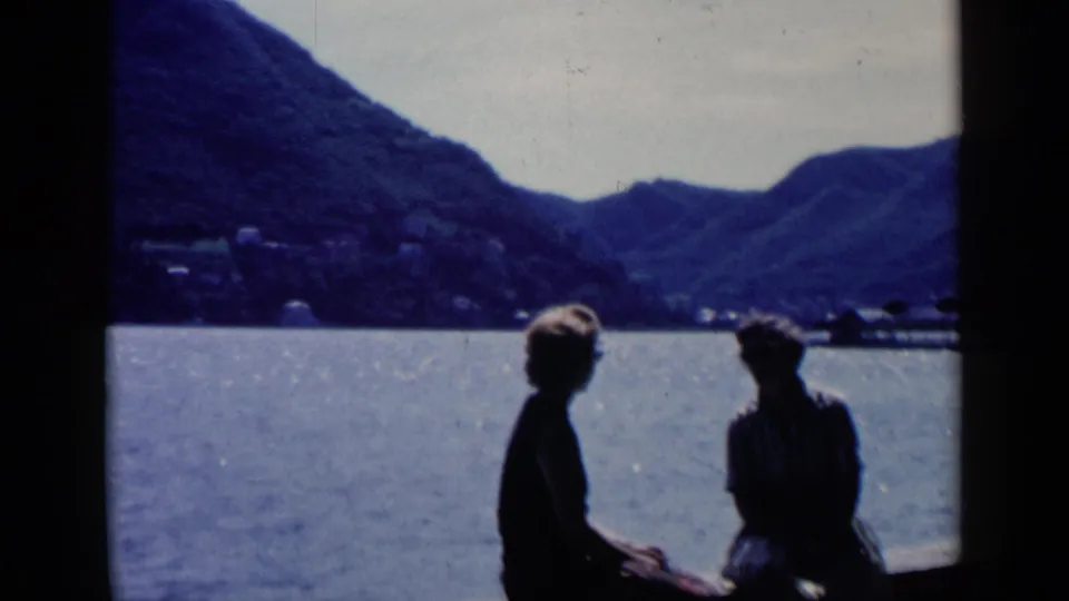 At the end of the clip, the couple remains seated by the lake in silhouette, with hills and shoreline structures behind them.