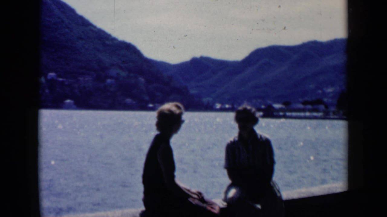 Two silhouetted figures sit beside bright water with mountains in the distance.