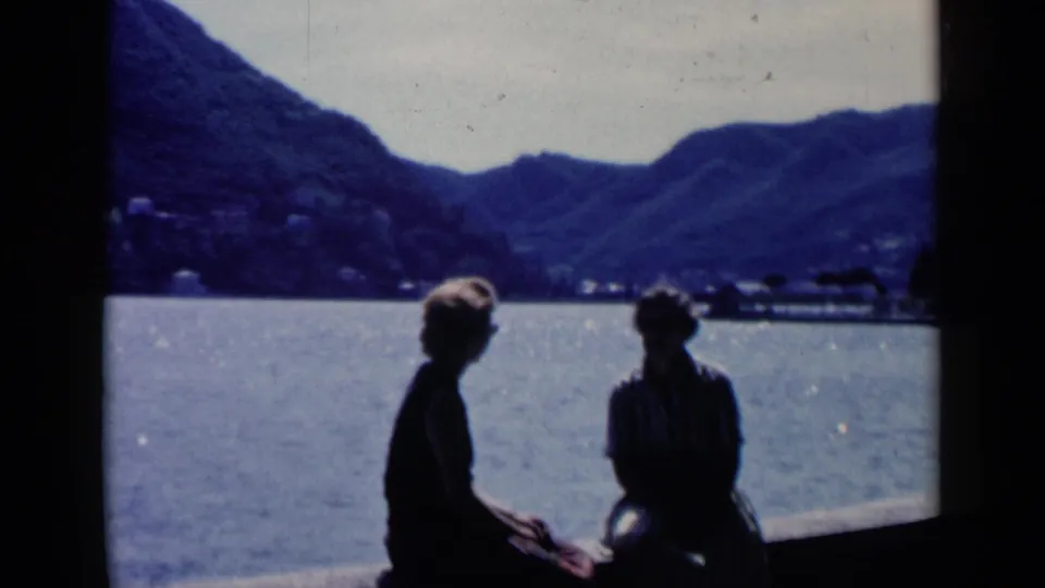 1960 8mm travel frame from Venice, Italy, showing two seated figures in silhouette against water and distant mountains.