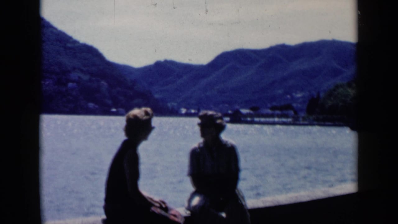 Two dark figures sit near the water with a mountainous shoreline beyond them.