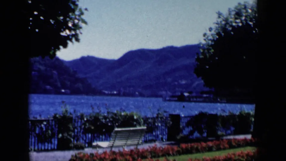 1960 8mm frame from Venice, Italy, showing a white garden bench, red flower bed, iron fence, water, and distant mountains.