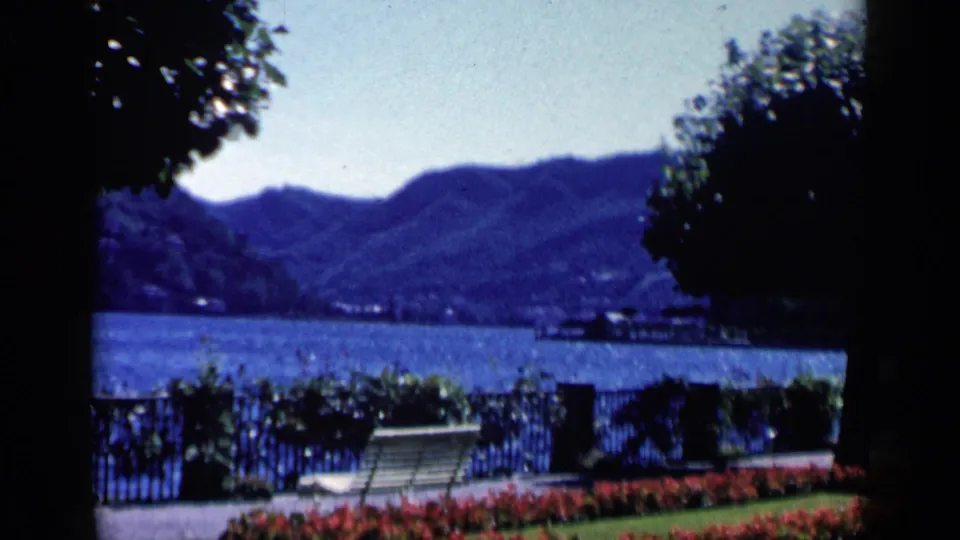 Soft 1960 8mm view in Venice, Italy, with a white bench, red flowers, dark foliage, iron fence, water, and mountains beyond.