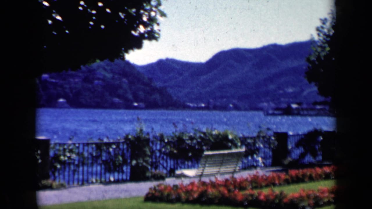 Cataloged 1960 Italy frame of a manicured lakeside garden with a bench, iron fence, blue water, and mountain horizon.