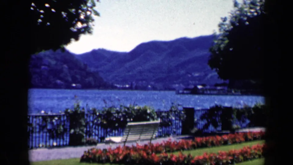 Cataloged 1960 Italy 8mm frame showing red flowers, a white bench, lakeside railing, blue water, and mountains.