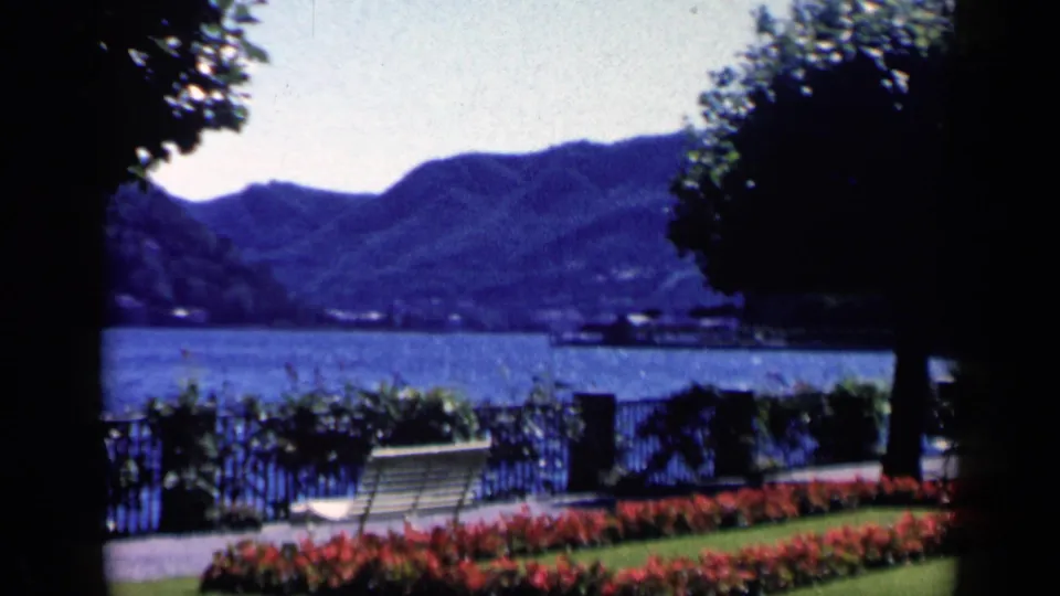 Cataloged 1960 Italy home-movie frame with tree shade over a lakeside garden, bench, railing, water, and mountains.