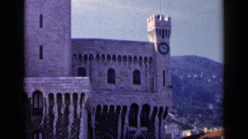 1960 8mm frame showing a European stone landmark with a prominent clock tower, arched openings, and soft hills behind the building.