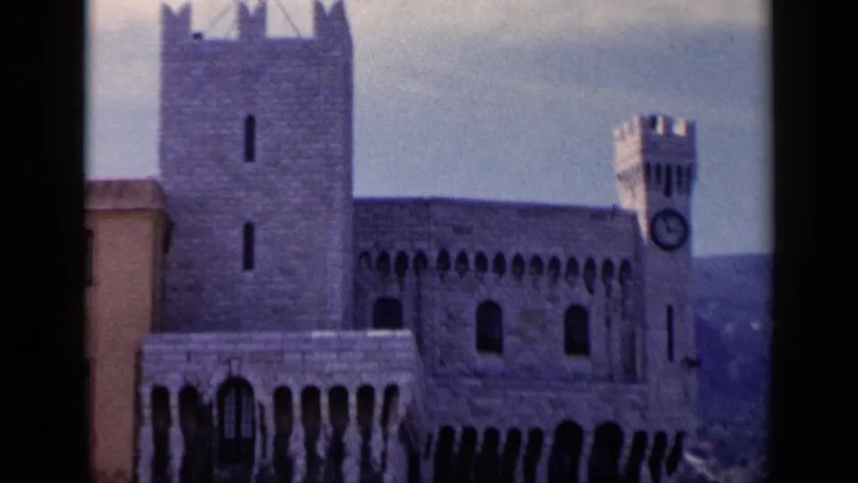 1960 8mm travel frame of the landmark from a slightly wider angle, showing a square tower, arched doorway, clock tower, and hillside setting.