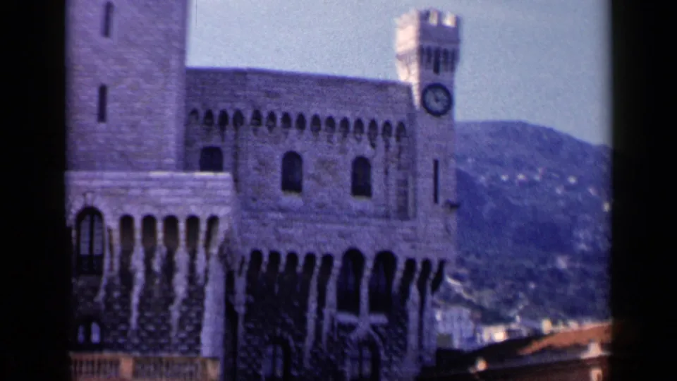 1960 8mm frame of the stone landmark in Italy, with the clock tower near the right side, arched windows across the facade, and hills beyond.