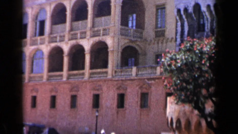1960 Venice travel frame showing arched colonnades, balcony railings, brick lower walls, and a flowering tree against the facade.