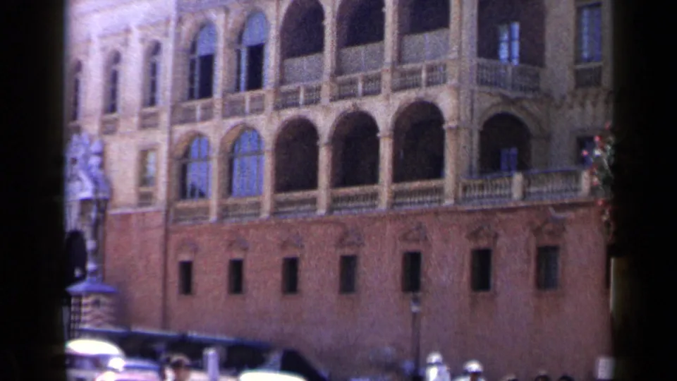 1960 Venice 8mm frame with arched windows, brown masonry, a partial statue at left, lamps, and tiny pedestrians near the bottom.