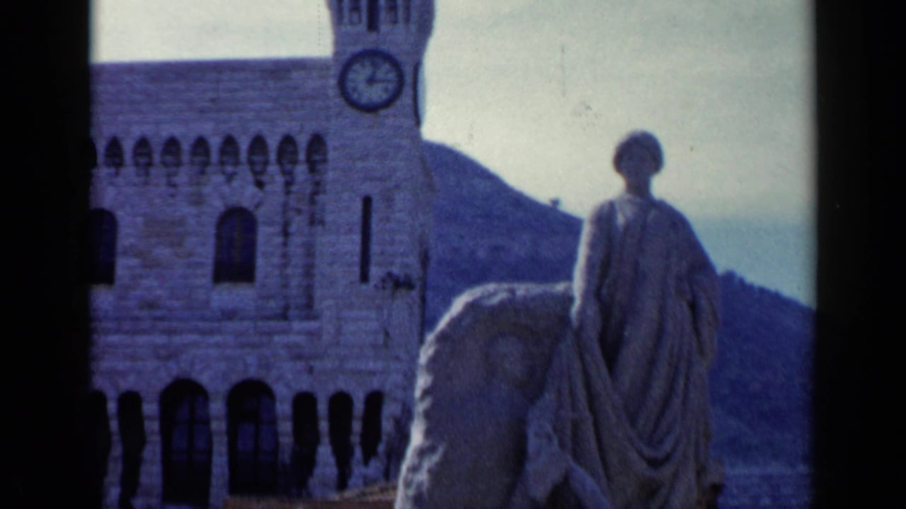 Stone statue before a clock tower and arched masonry with a hillside behind.